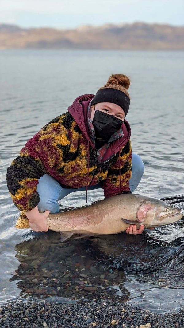 Angler in a mask and beanie holding a large Cutthroat Trout in shallow water.