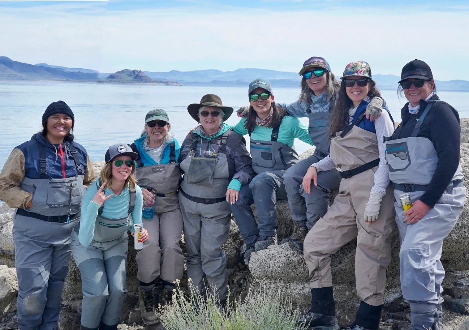 Group of anglers in waders posing on a rocky shoreline by a large lake.