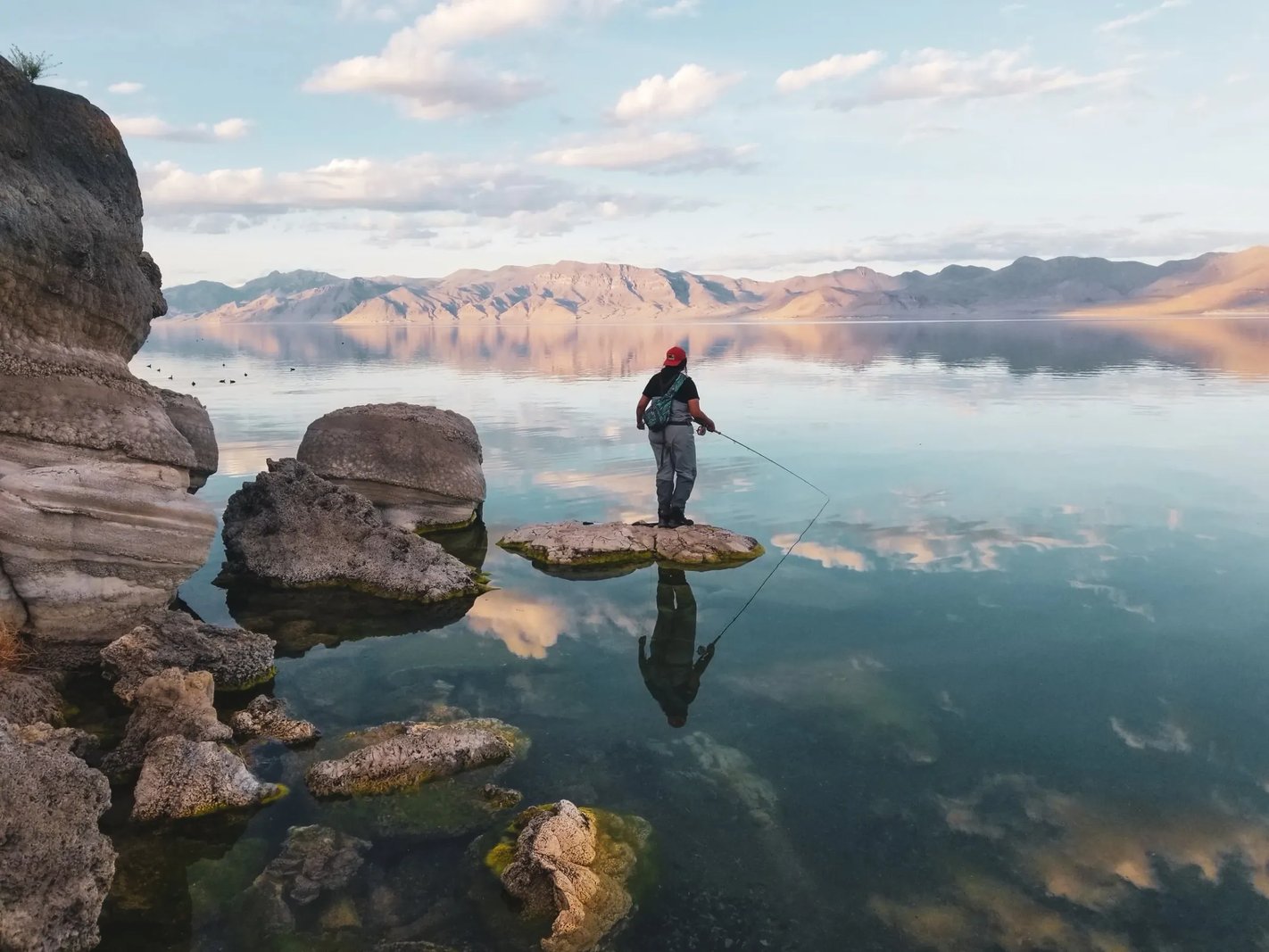 Angler standing on a rock in the water, fly fishing with a scenic lake and mountains.