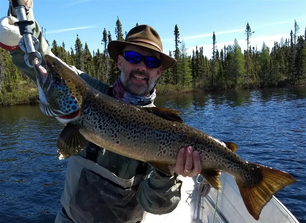 Man in a hat and sunglasses holding a large speckled fish with a fish gripper.