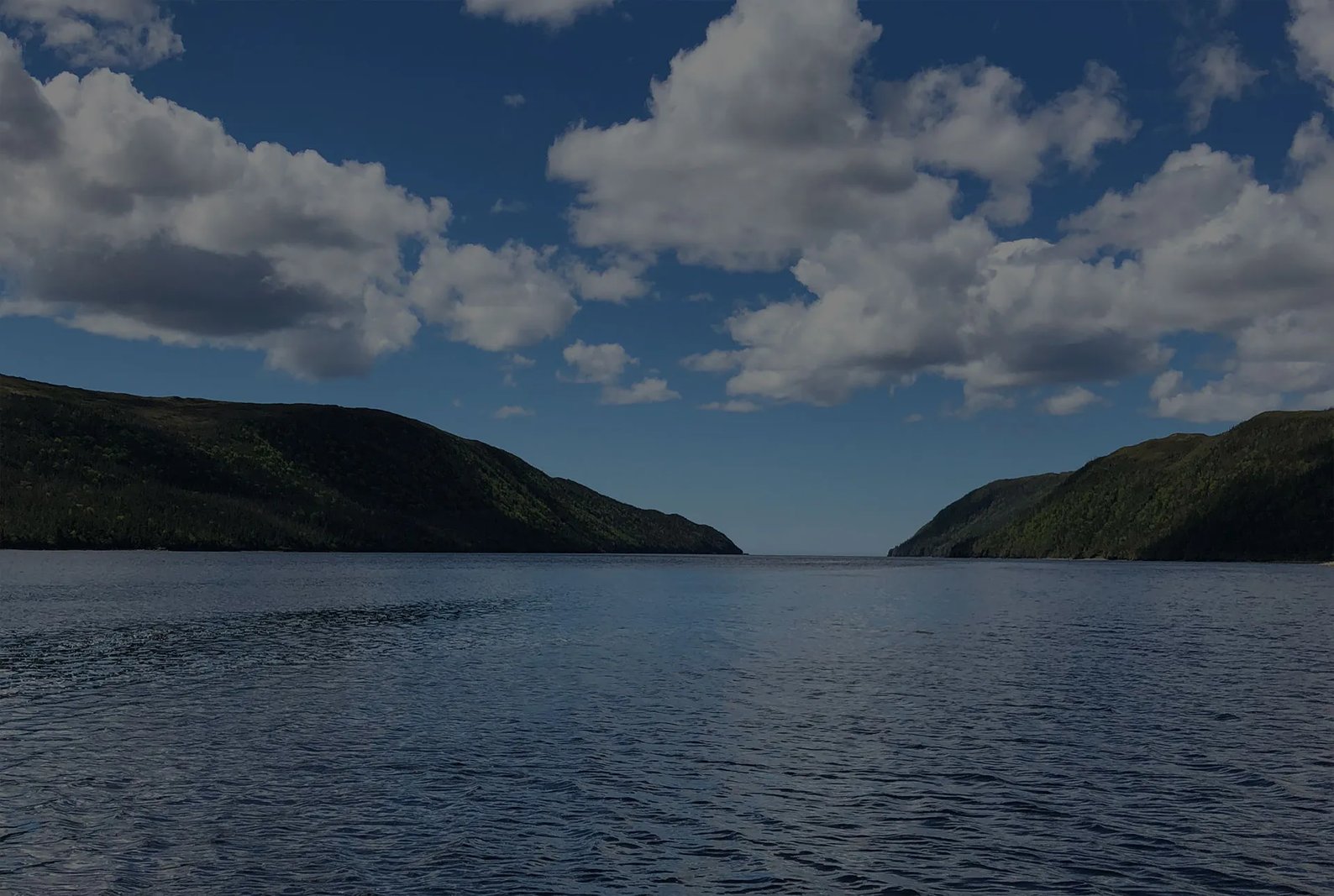 Wide view of a calm bay between two green mountains under a cloudy sky.