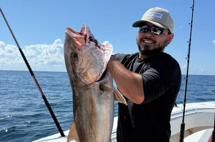 Man in sunglasses and a cap holding a large Amberjack on a boat under a sunny sky