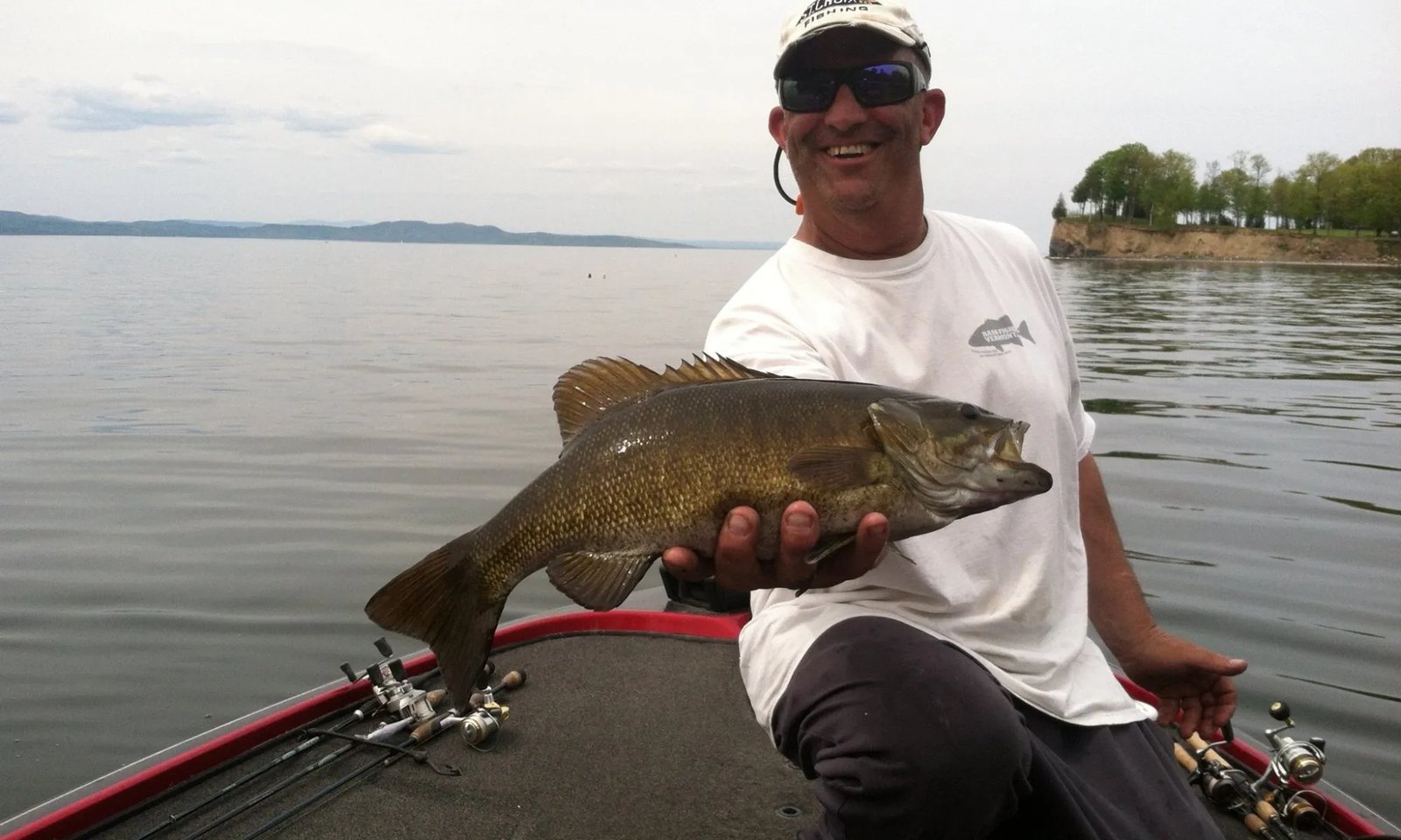 Angler on a bass boat holding a large Smallmouth Bass with a lake and distant shore