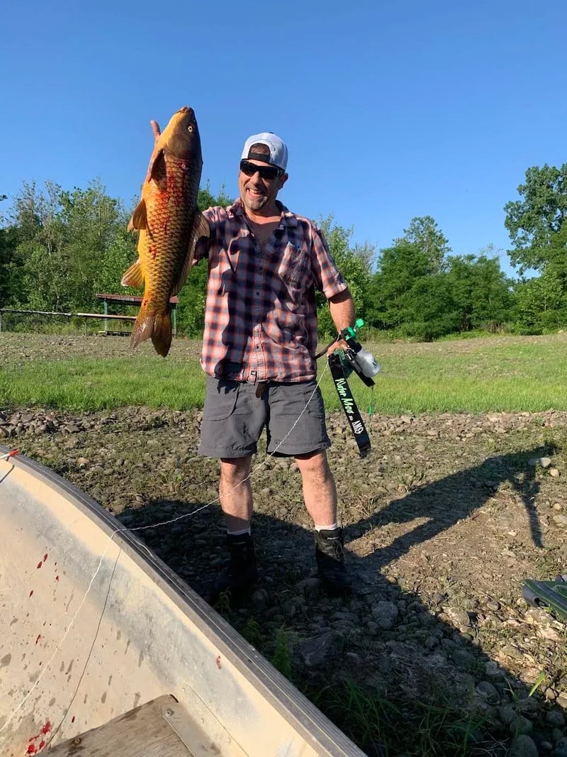 Man in a plaid shirt holding a large golden Common Carp with bowfishing gear