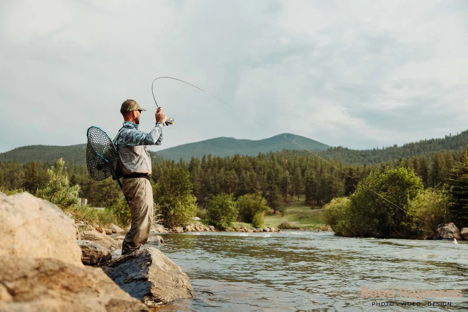 Angler casting a fly rod from a rocky river bank with forested mountains in the background