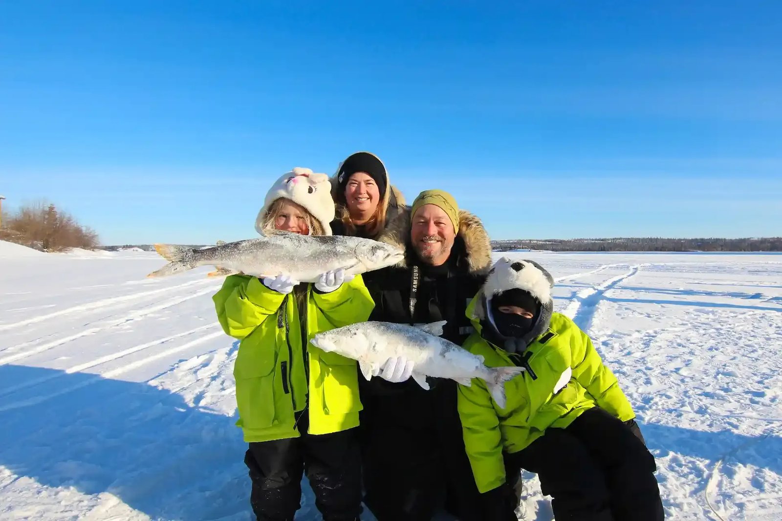 Group of anglers in winter gear holding large Lake Trout on a snowy lake.