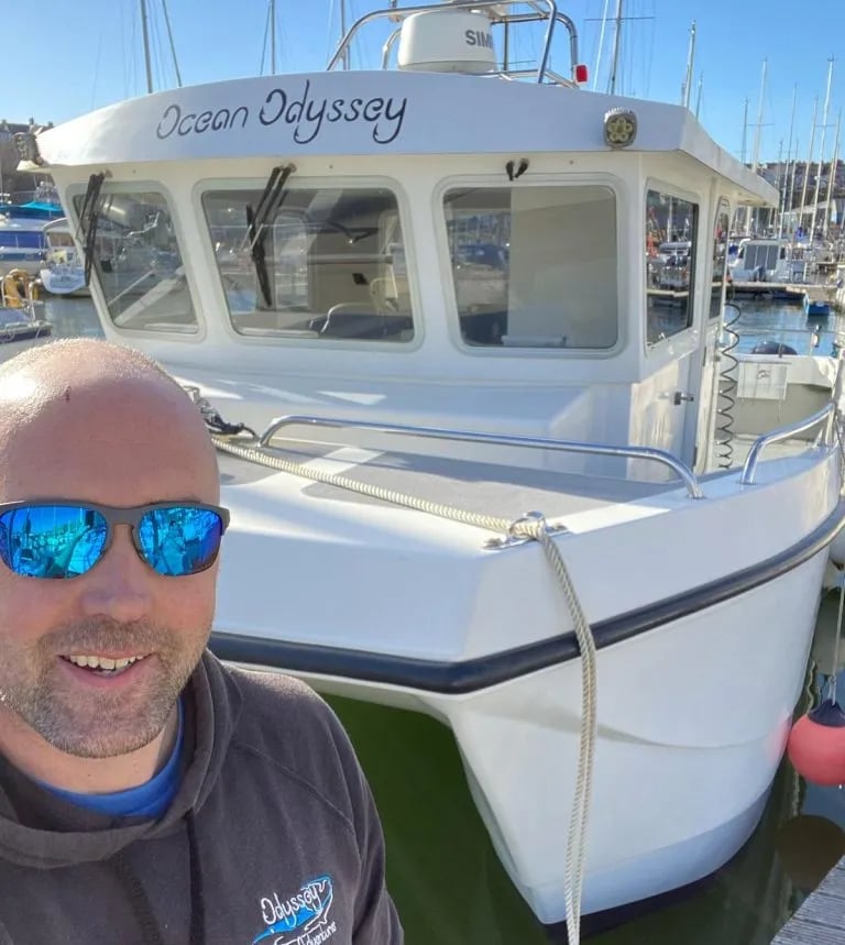 Man in sunglasses smiling in front of the 'Ocean Odyssey' boat at the dock.