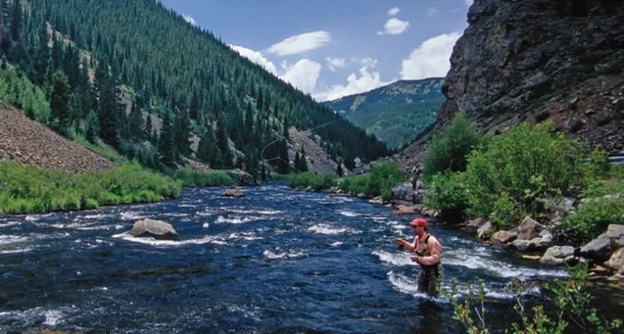 Angler wading in a wide mountain river surrounded by steep, forested valleys.