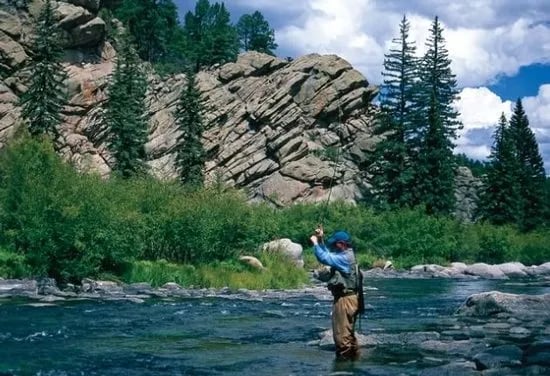 Angler casting a fly rod in a river with large rocky formations and pine trees.