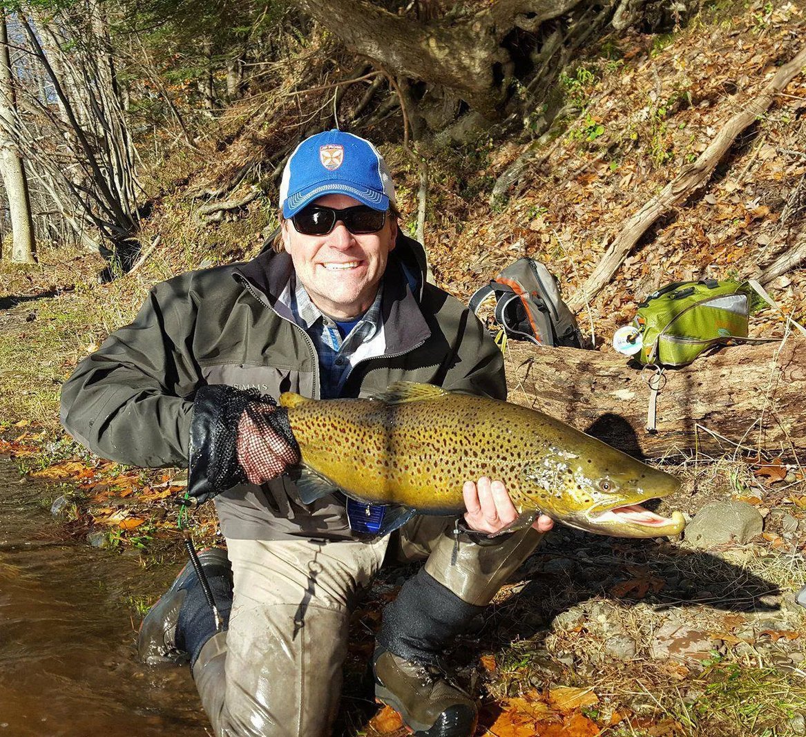 Smiling angler holding a large Brown Trout in a river with green trees