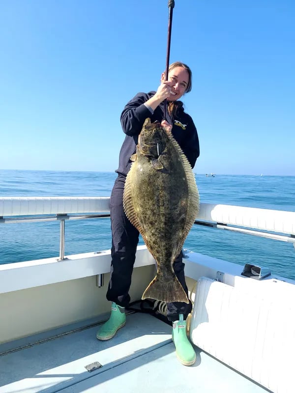 Angler on a boat holding a large Halibut by the mouth against a clear blue sky