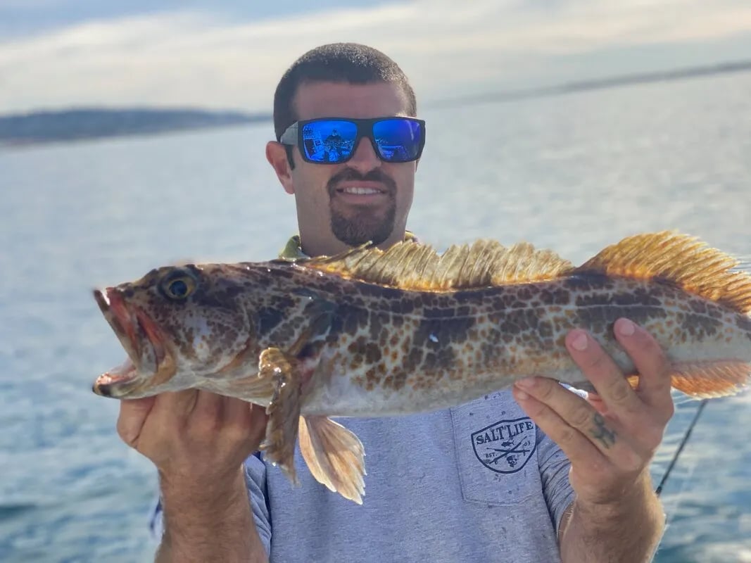 Angler in sunglasses holding a Lingcod with distinctive markings on a boat