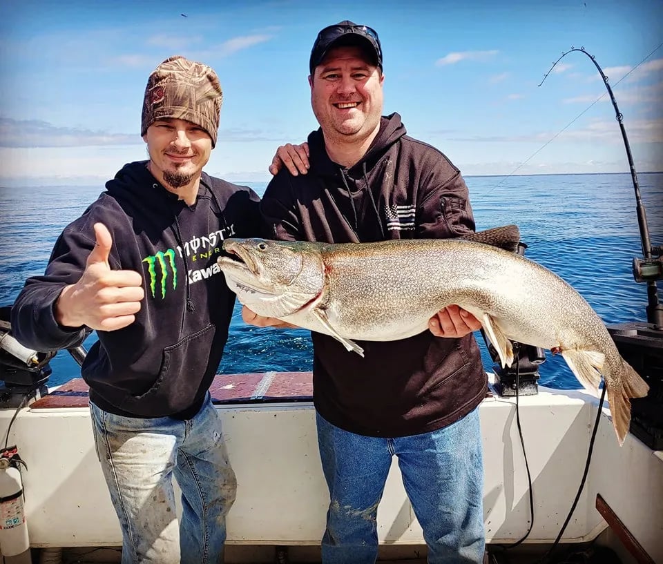 Two anglers holding a large Lake Trout on a boat, giving a thumbs up