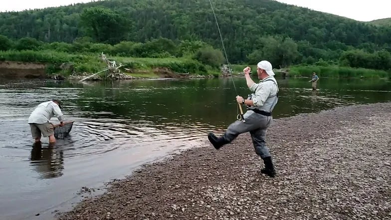 Two anglers on a rocky river bank, one celebrating a catch, the other in the water with a net.