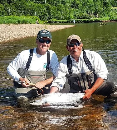 Two smiling anglers kneeling in a river, holding a large fish.