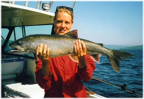 Woman in a red shirt holding a large Lake Trout on a boat with blue water and mountains