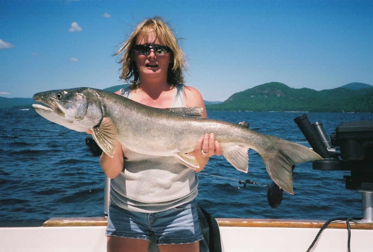 Woman in sunglasses holding a very large Lake Trout on a boat under a bright blue sky
