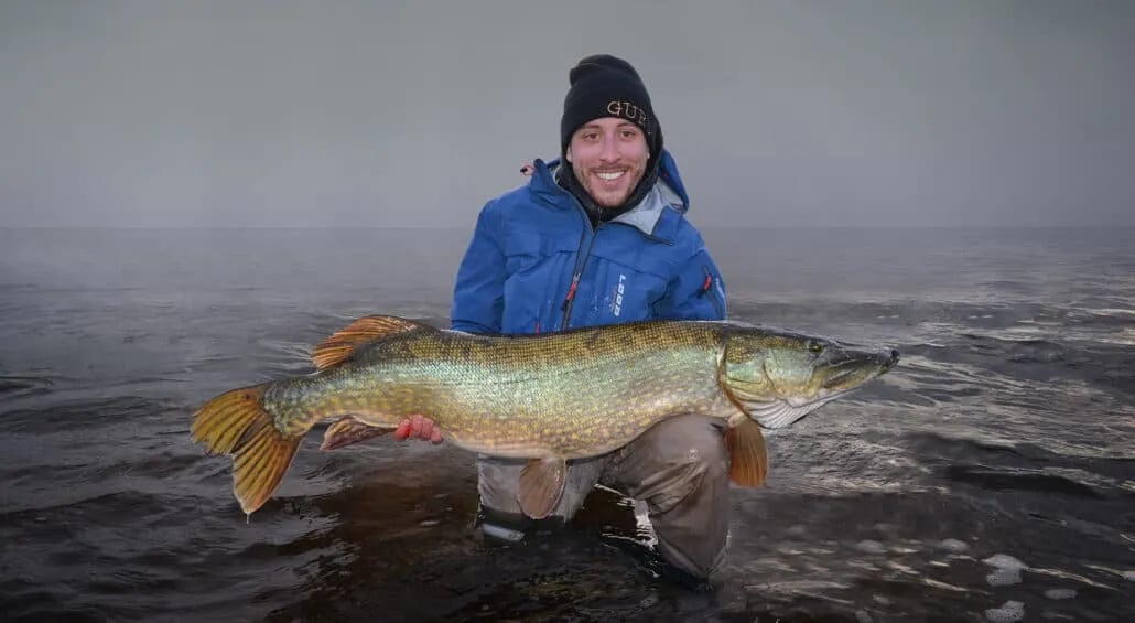 Angler wading in a wide river under a cloudy sky with forested banks