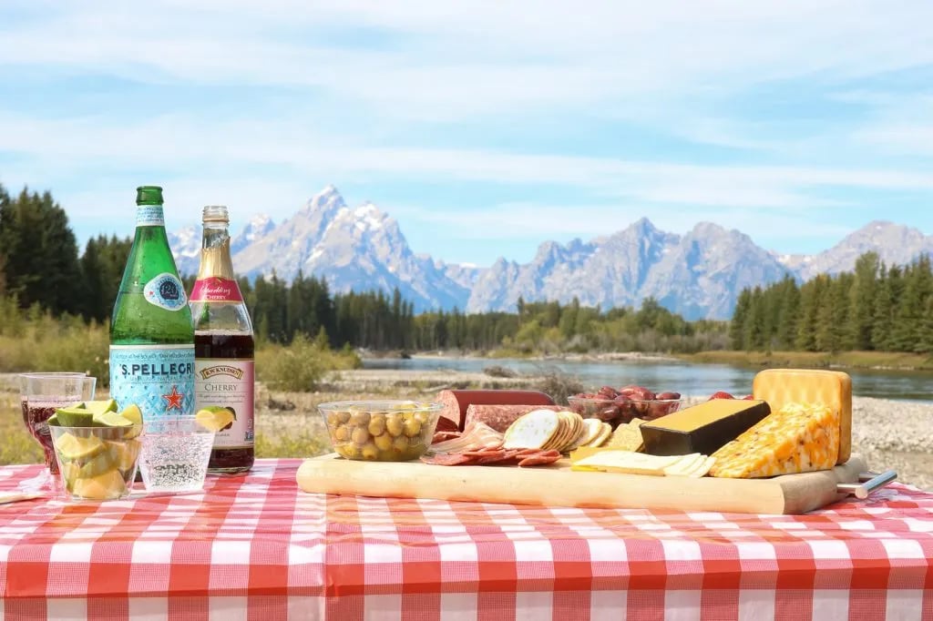Picnic spread on a red and white checkered tablecloth with a river and Grand Teton mountains in the background