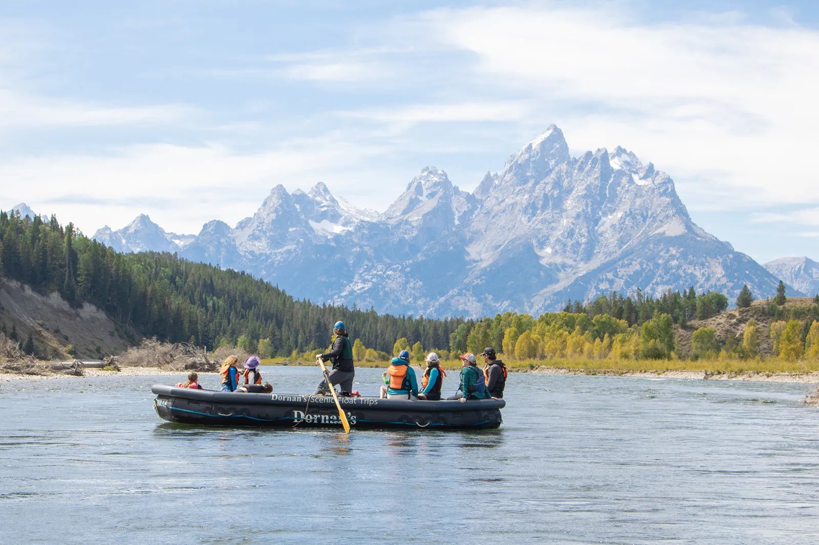 Guide and passengers in a raft on a river with a dense forest and the Grand Teton mountains