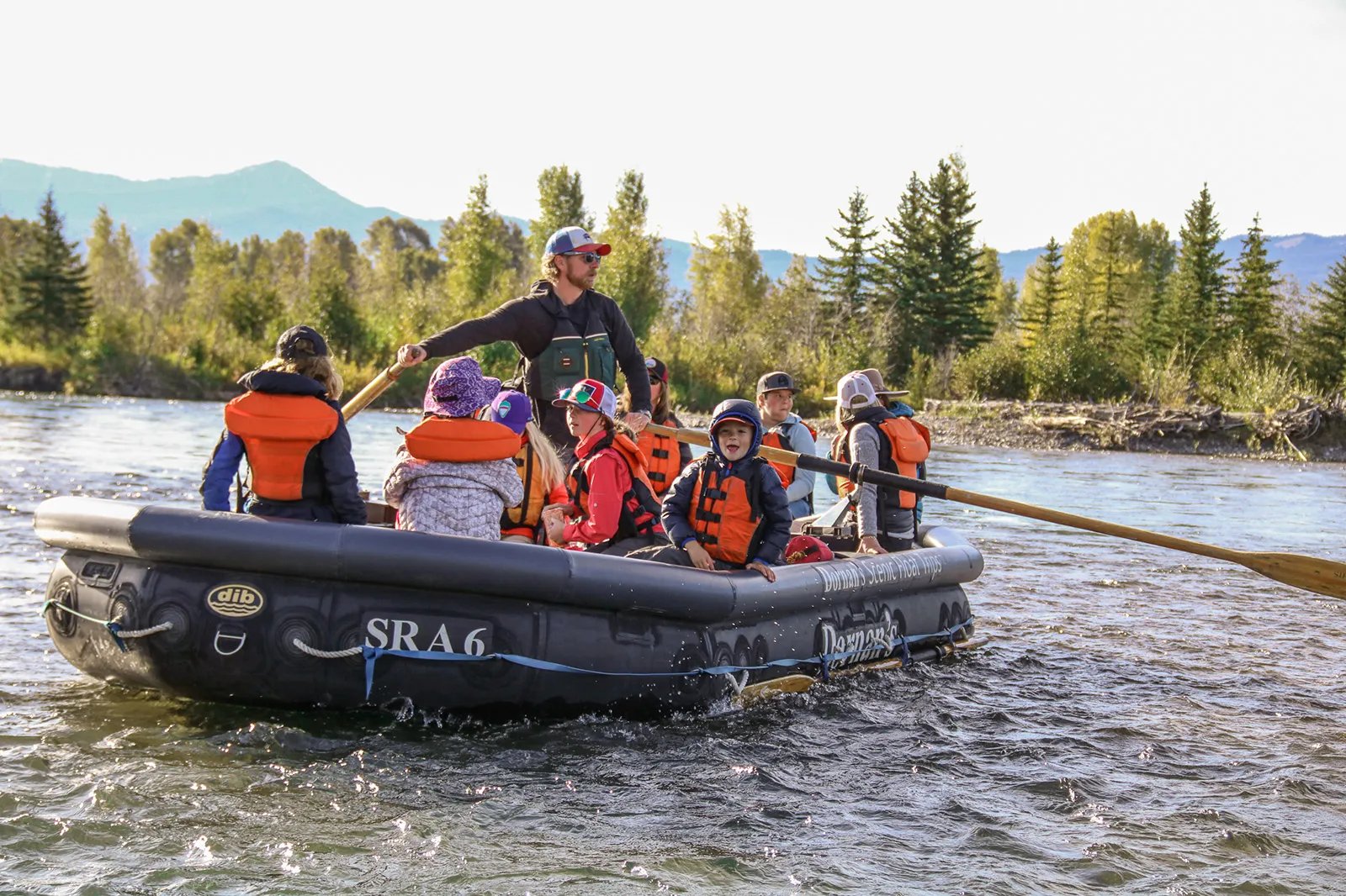 Guide poling a raft with several passengers wearing life jackets on a sunny river