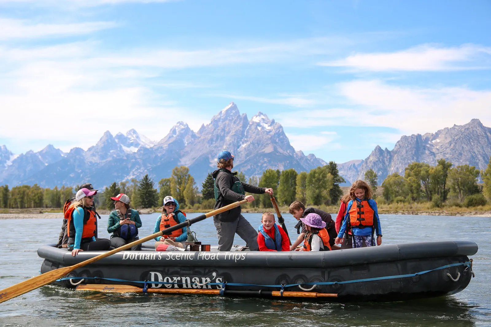 Guide poling a raft full of passengers down a river with the snow-capped Grand Teton mountains behind them