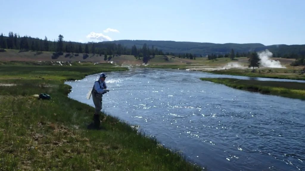 Angler wading and fly fishing in a wide, clear river with distant hills and steam vents