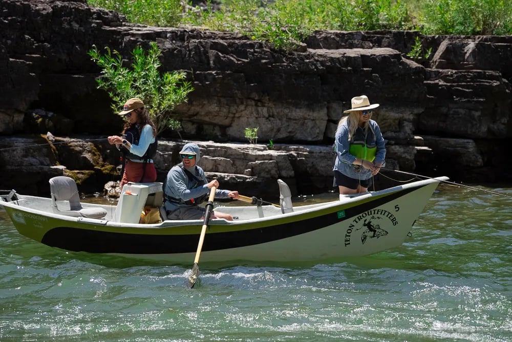Drift boat with two anglers and a guide rowing on a river with rocky banks.