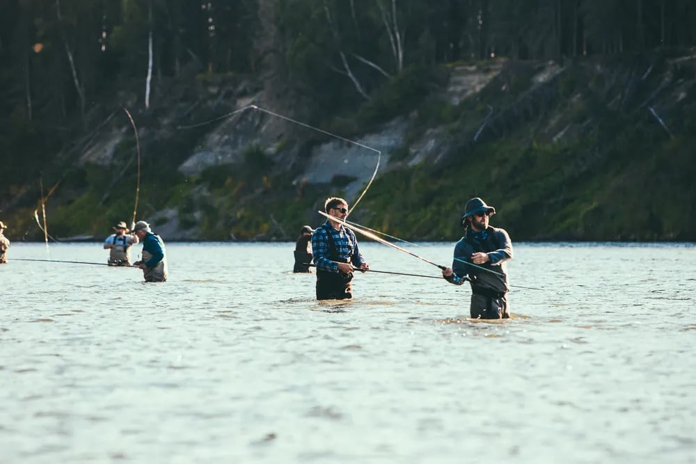 Multiple anglers wading and casting in a wide river with a forested bank.
