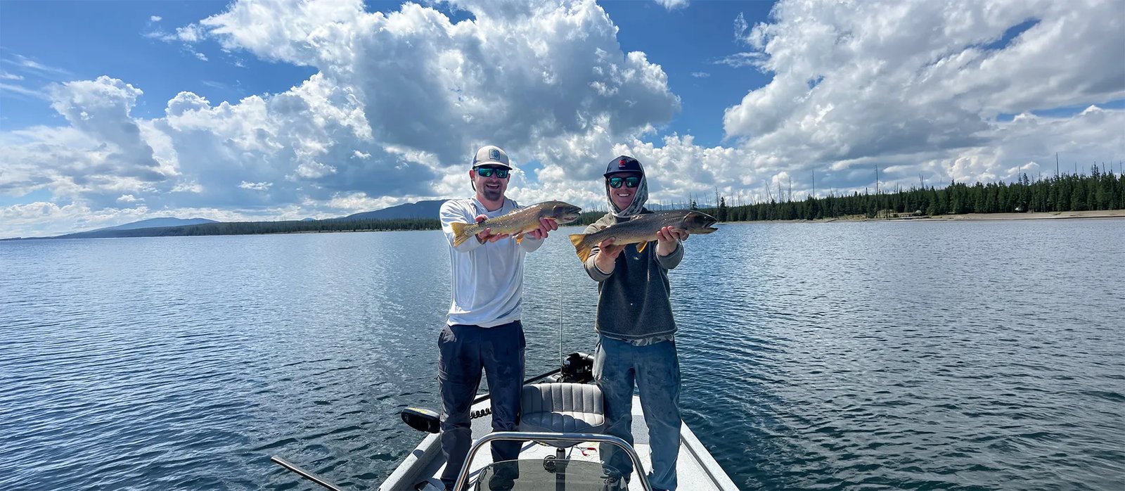 Two anglers in a boat holding up Brown Trout on a large lake under a cloudy sky.