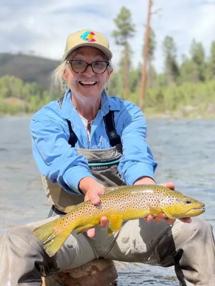 Woman in waders smiling, holding a spotted Brown Trout in a river.