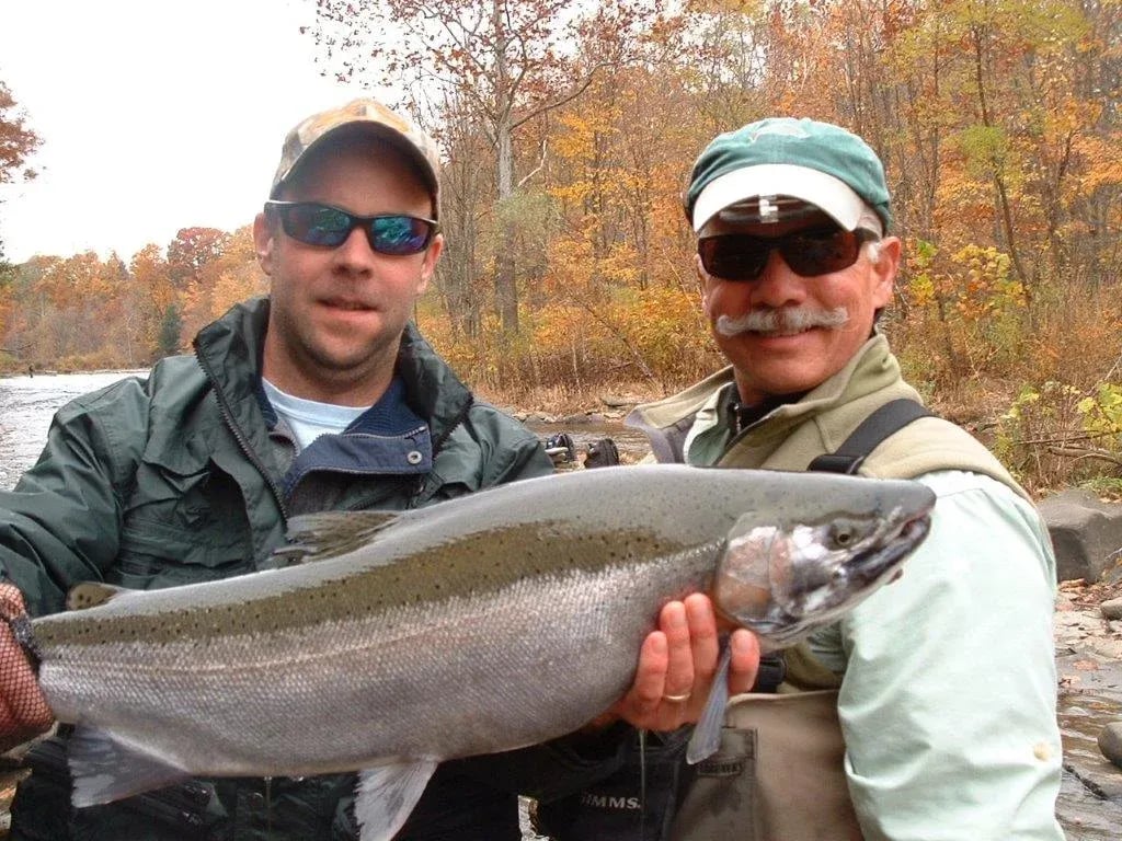 Two anglers in a drift boat holding a large Steelhead on a river with bare trees