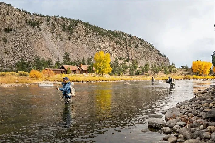 Two anglers wading and fly fishing in a river with fall foliage and mountains