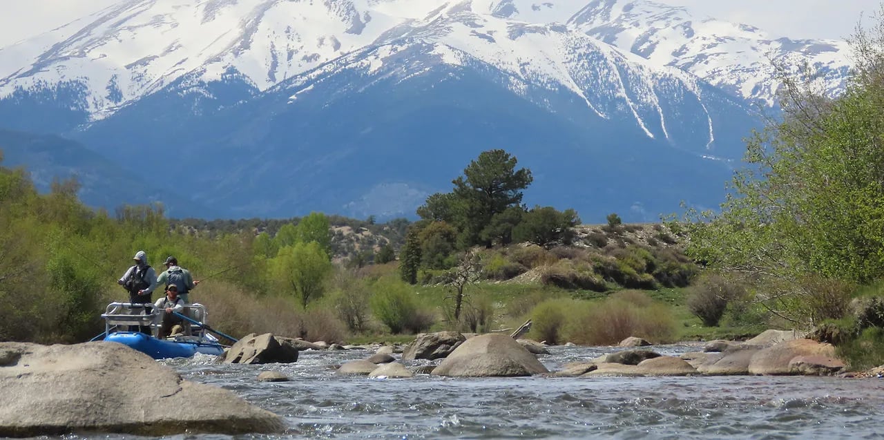 Two anglers in a raft on a river with a snowy mountain in the background