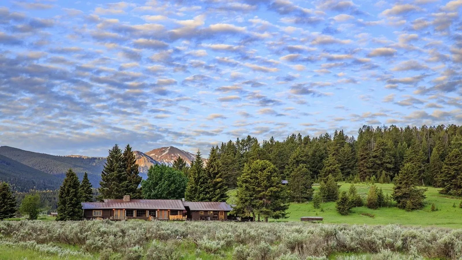 Lodge building nestled in a green valley surrounded by pine trees and mountains.
