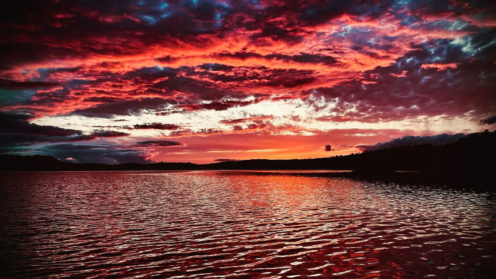 Dramatic red and orange sunset over a calm lake with dark silhouettes of distant trees