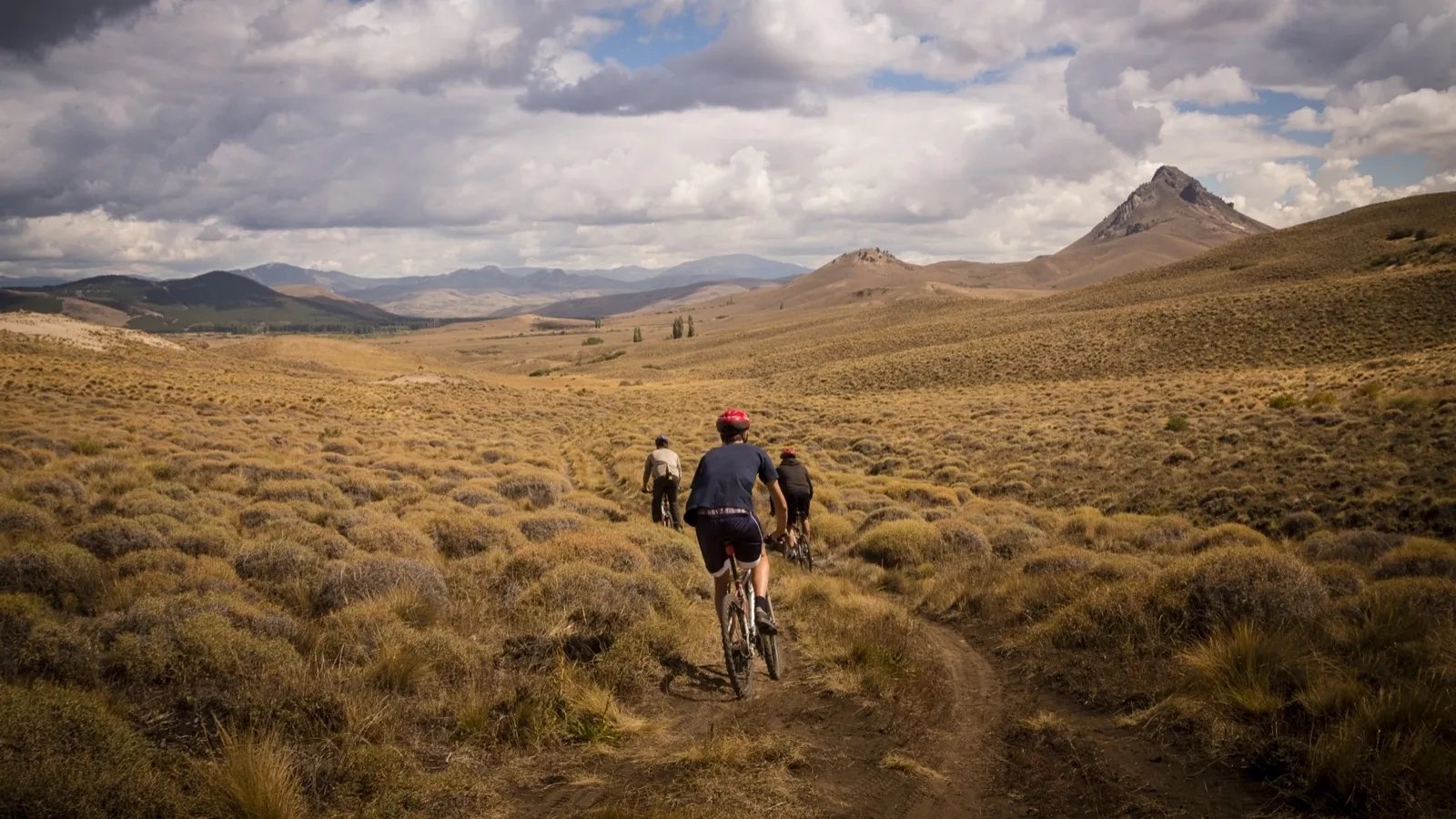 Mountain biker riding away on a trail through a vast, dry grassy landscape with a mountain peak in the distance