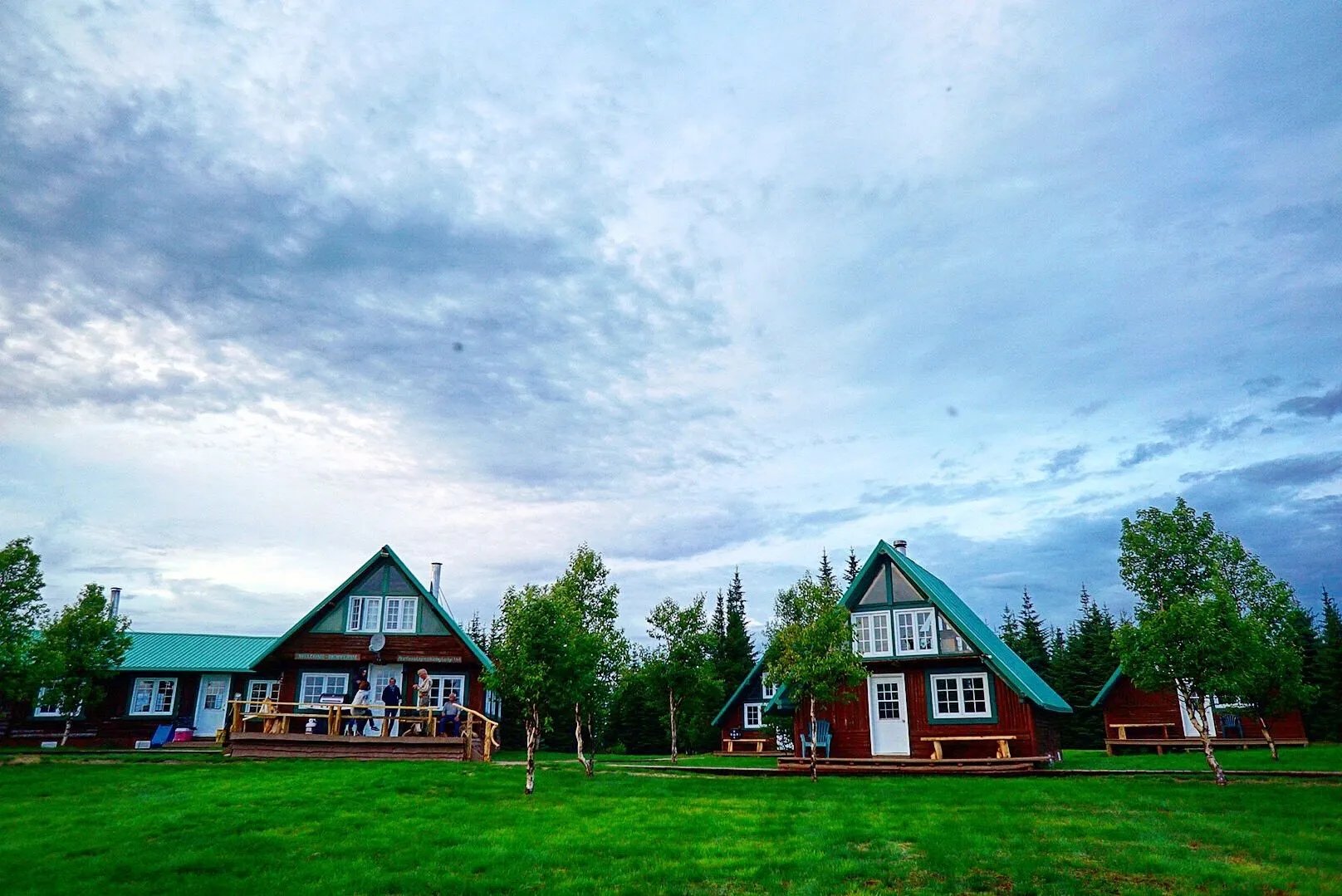 Several A-frame cabins with green roofs on a grassy field under a cloudy sky