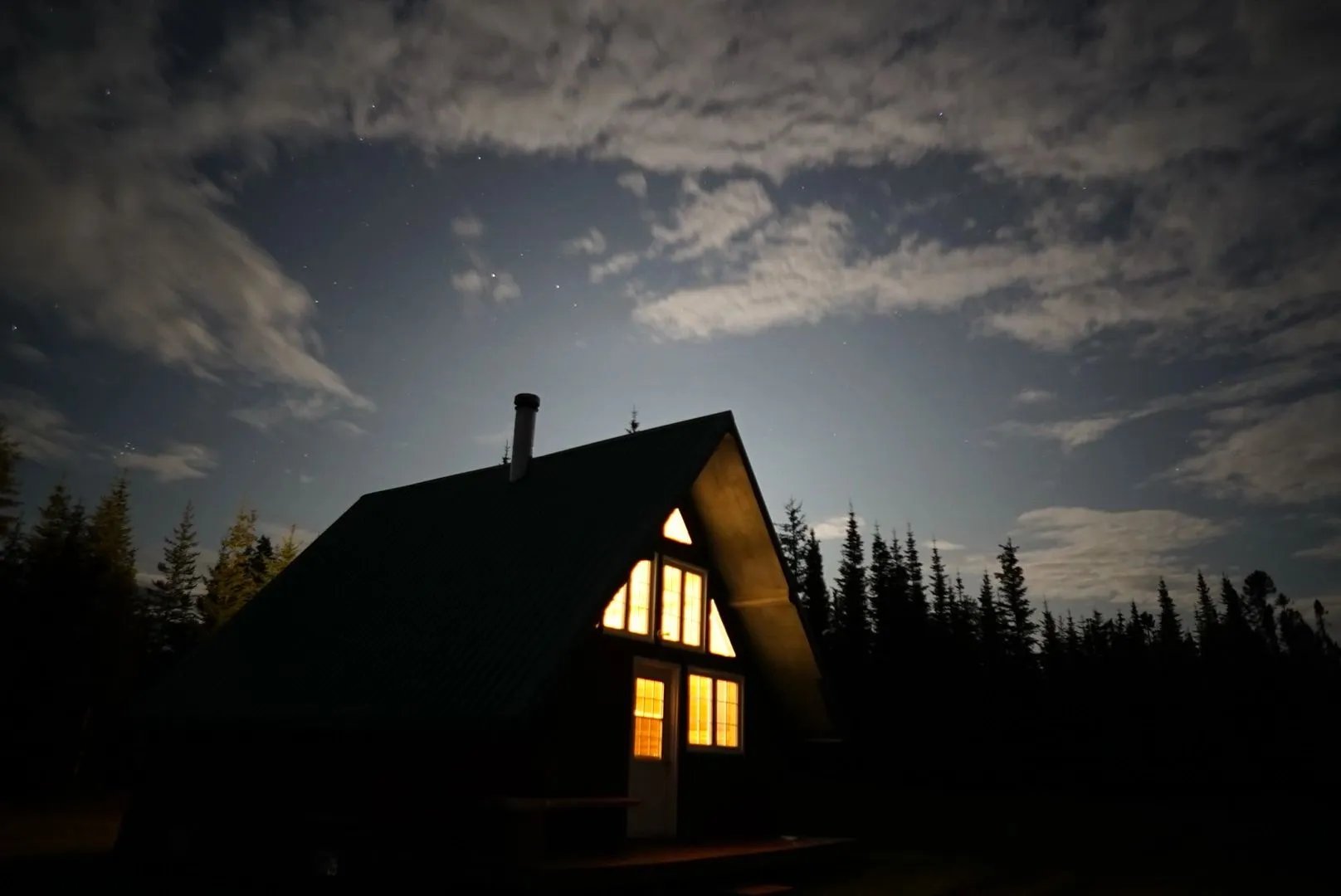 A-frame cabin at night with warm light glowing from windows, silhouetted against a cloudy sky