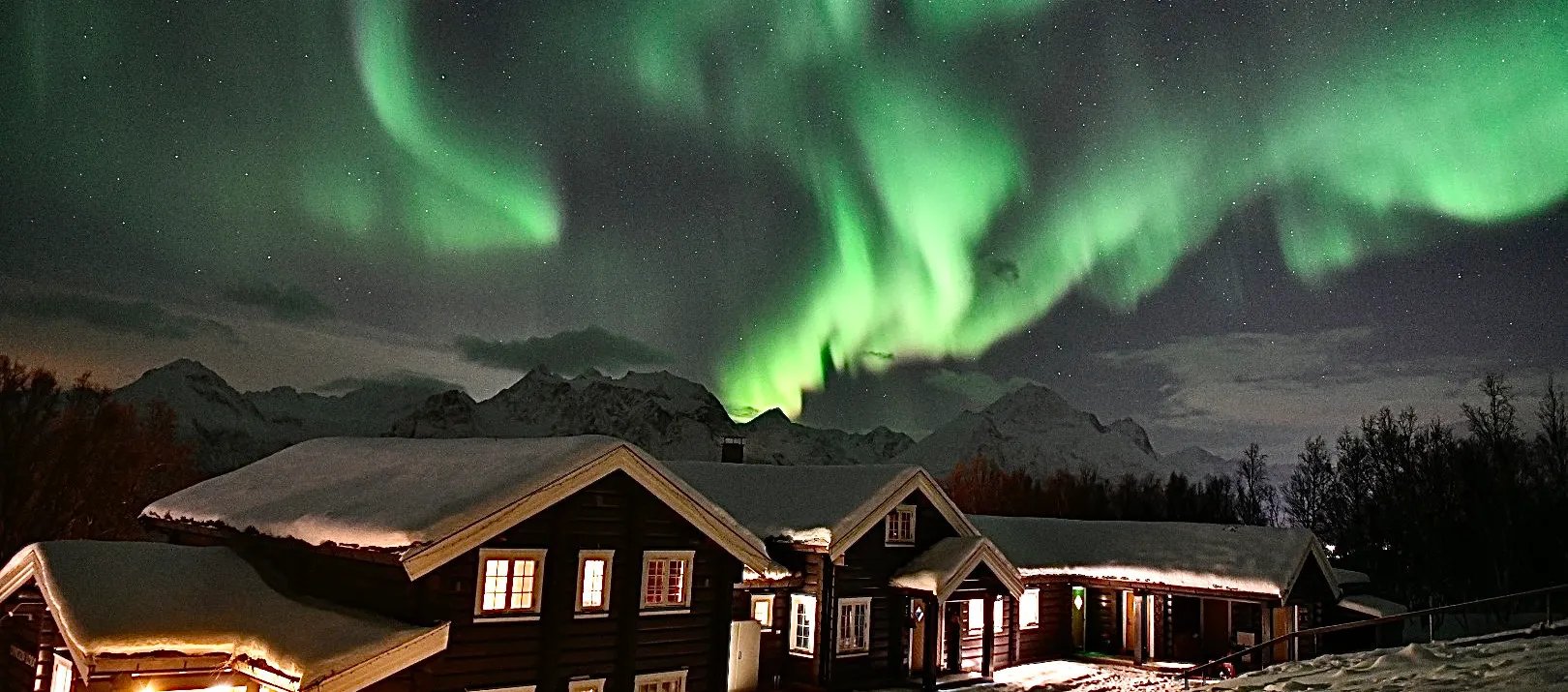 Snow-covered lodge buildings illuminated at night under a spectacular green aurora borealis