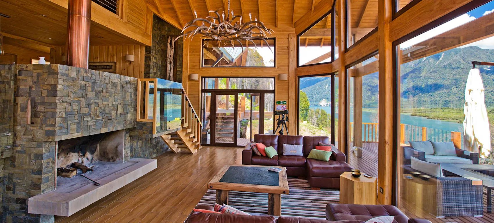 Spacious lodge living room with stone fireplace, wooden beams, and large windows overlooking a lake
