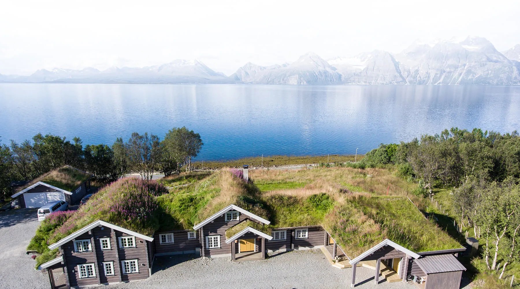 Aerial view of Lyngen Lodge, a complex of grass-roofed buildings nestled by a calm fjord with mountains