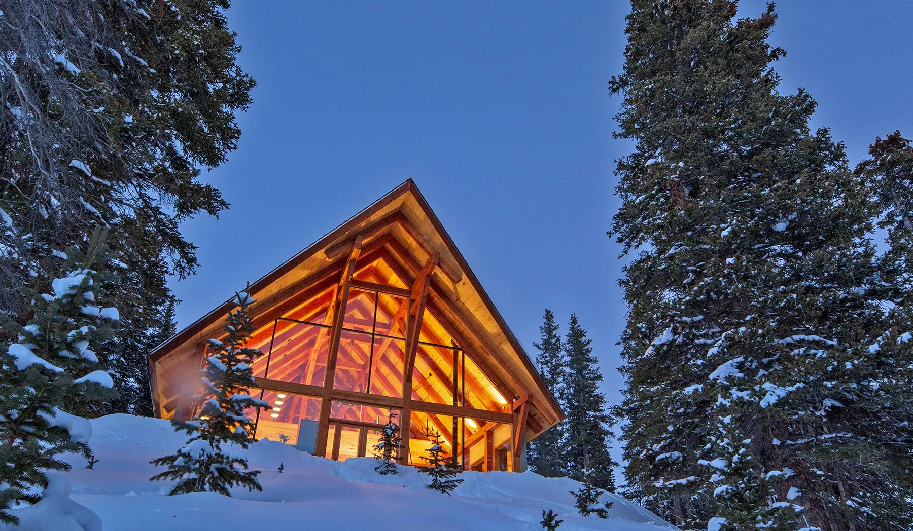 A-frame lodge exterior glowing with warm light at dusk, surrounded by snowy pines