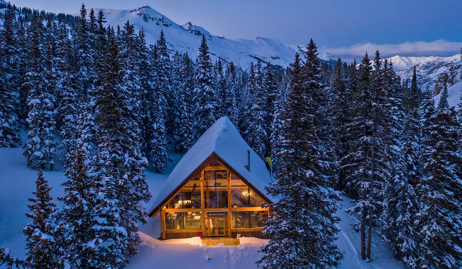 A-frame lodge glowing with warm light at dusk, nestled in a snowy forest with mountains in the distance