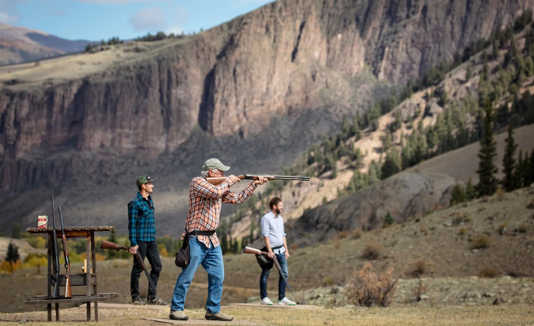Man shooting a shotgun at sporting clays in a scenic mountain valley with other participants.