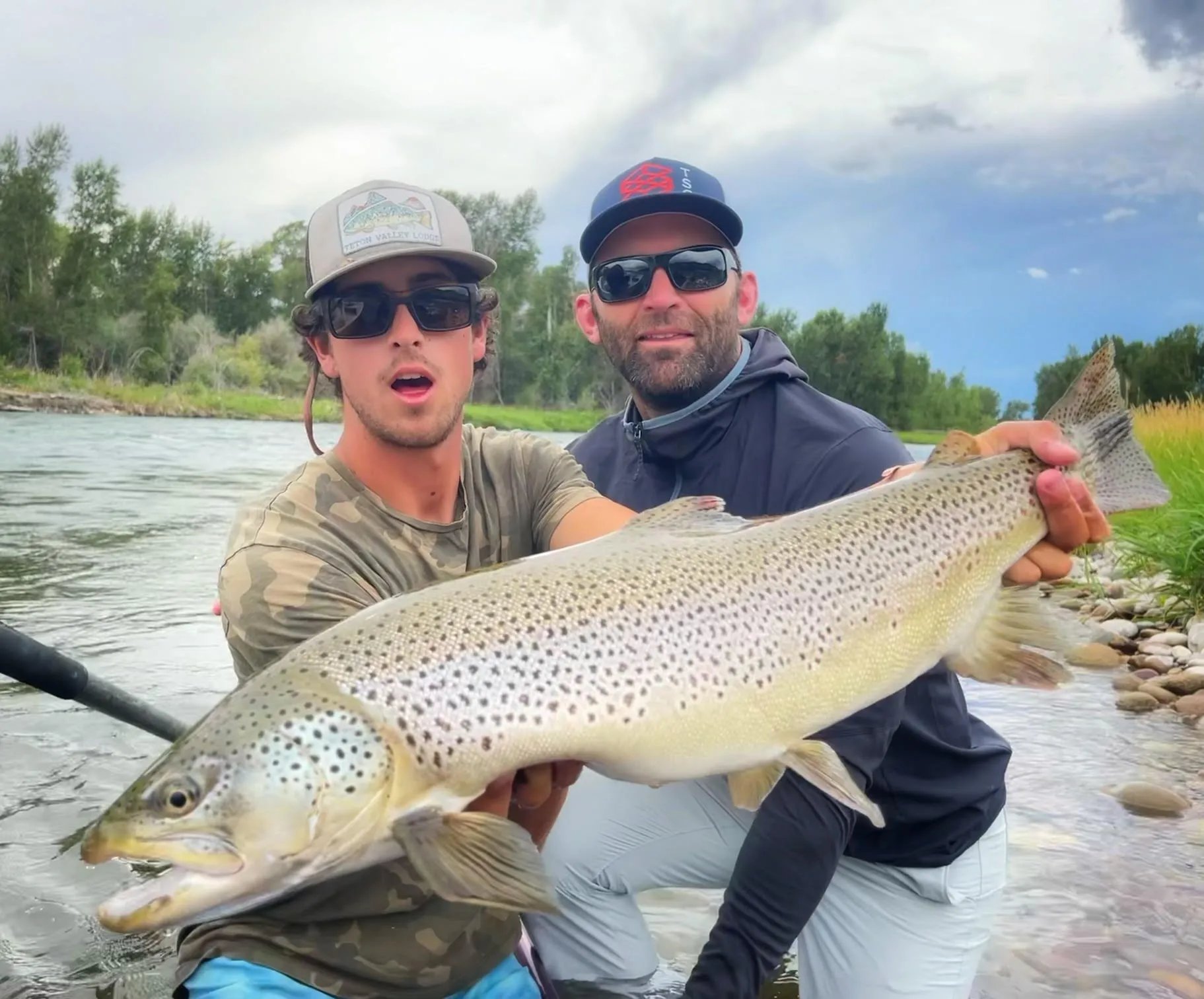 Two smiling anglers holding a very large brown trout in a river