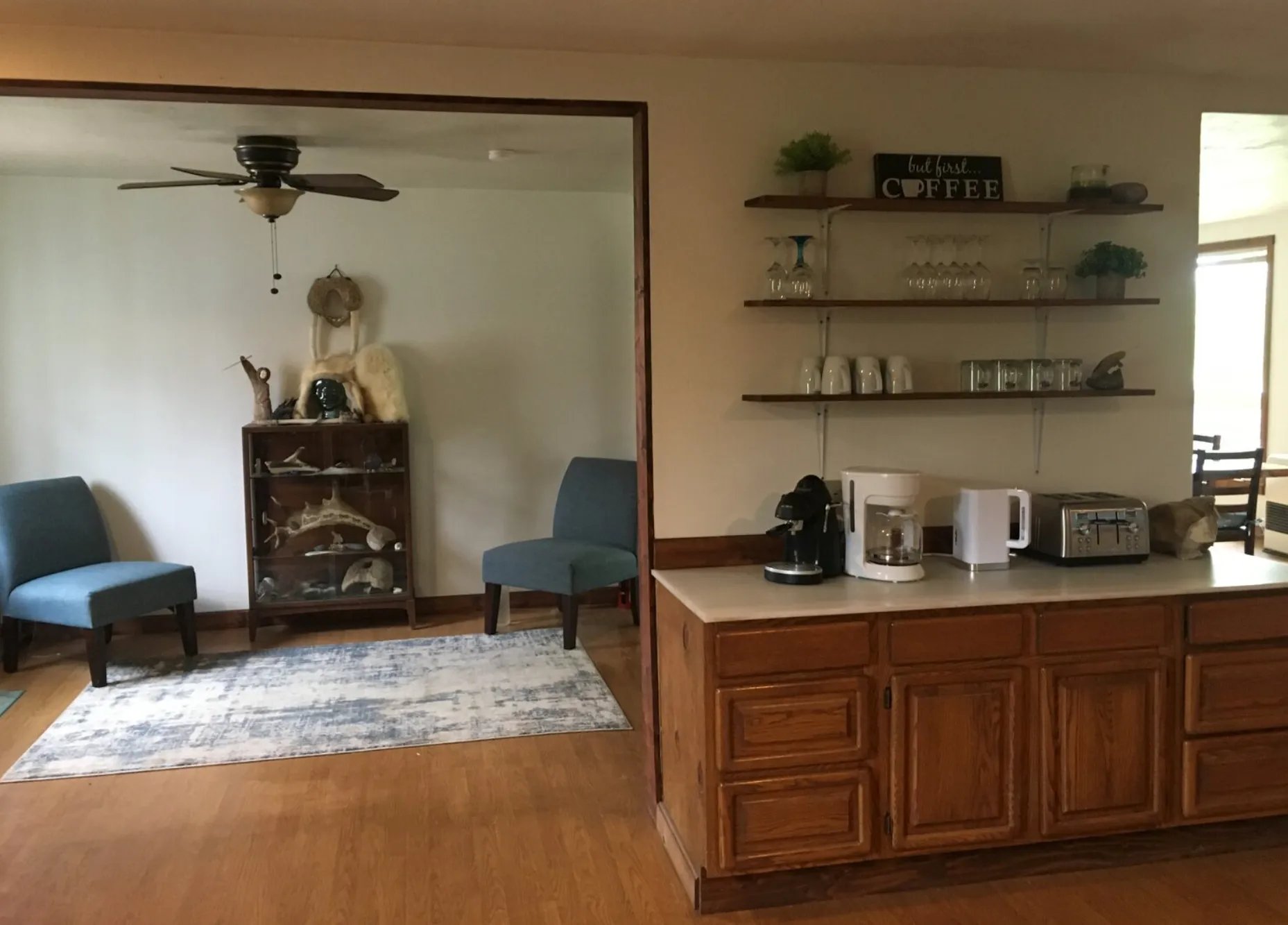 Interior view of a living space featuring blue chairs, a glass cabinet with decorative items, a coffee station with coffee makers and a toaster, and wooden shelves holding glasses and cups, under a sign reading 'but first... coffee.'"