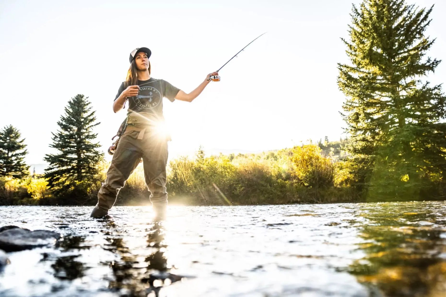 Woman fly fishing in a sunlit river with pine trees