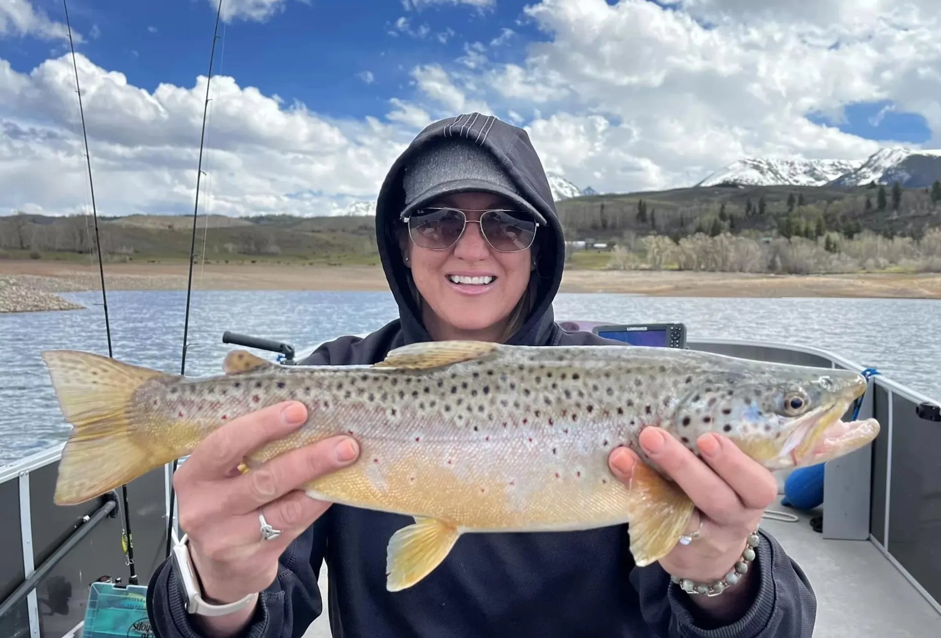 Woman in a hoodie holding a large brown trout on a boat
