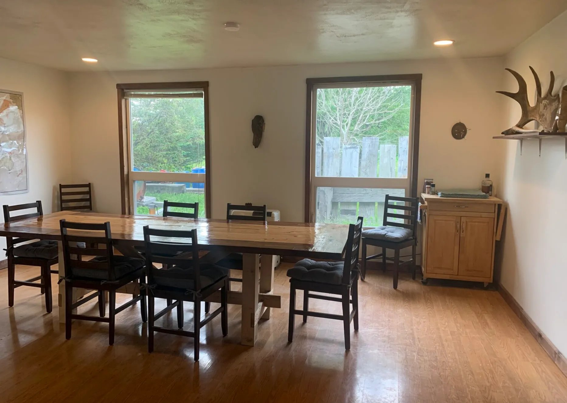 Dining room with wooden table, eight chairs, wall decor, two large windows, wooden floor, antler decoration, and a side cabinet.